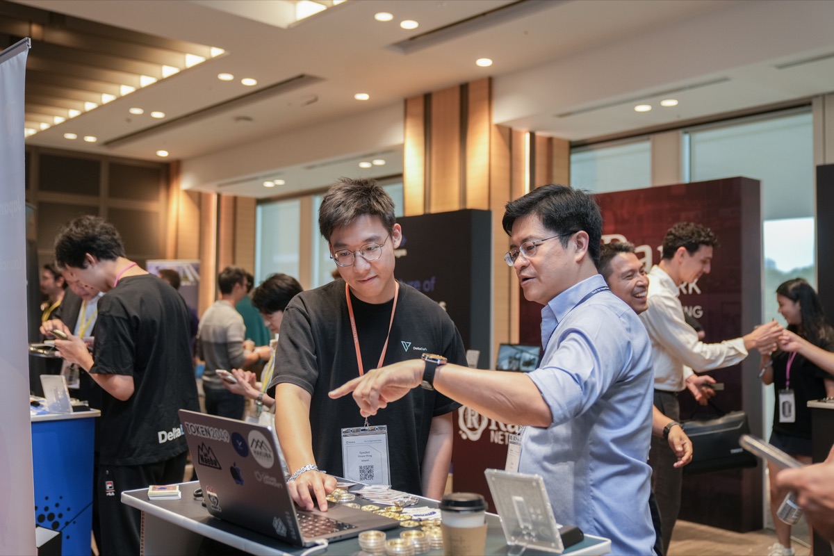 Two attendees in event lanyards working over open laptops at an exhibition booth, with other participants networking between booths in a bright conference hall